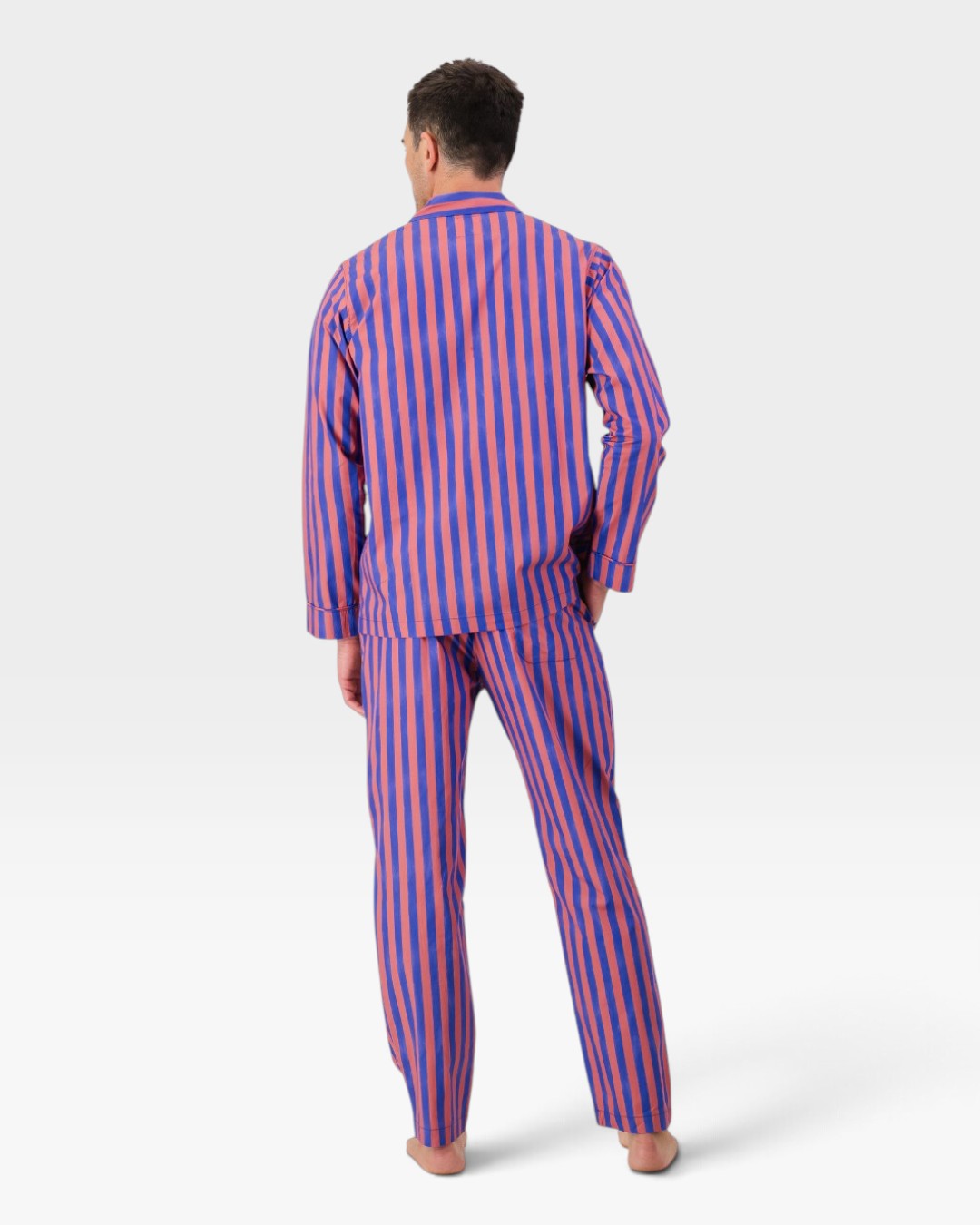 A man viewed from behind wearing a matching long-sleeve button-up pyjama shirt and trousers set with vertical red and blue stripes, on a light gray studio background.