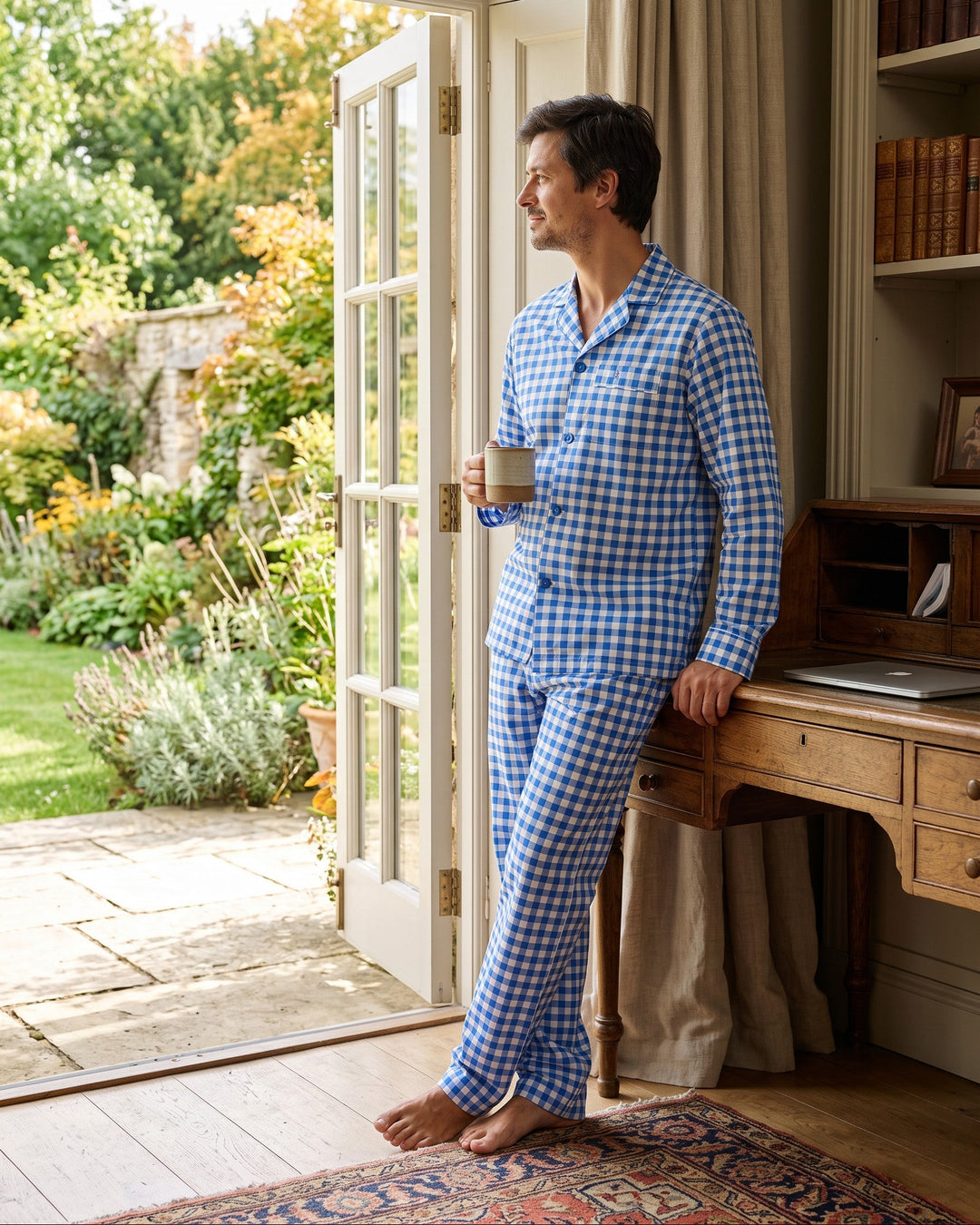 A man wearing blue and white checkered pyjamas stands barefoot in a room, holding a mug and looking out an open door to a garden patio.